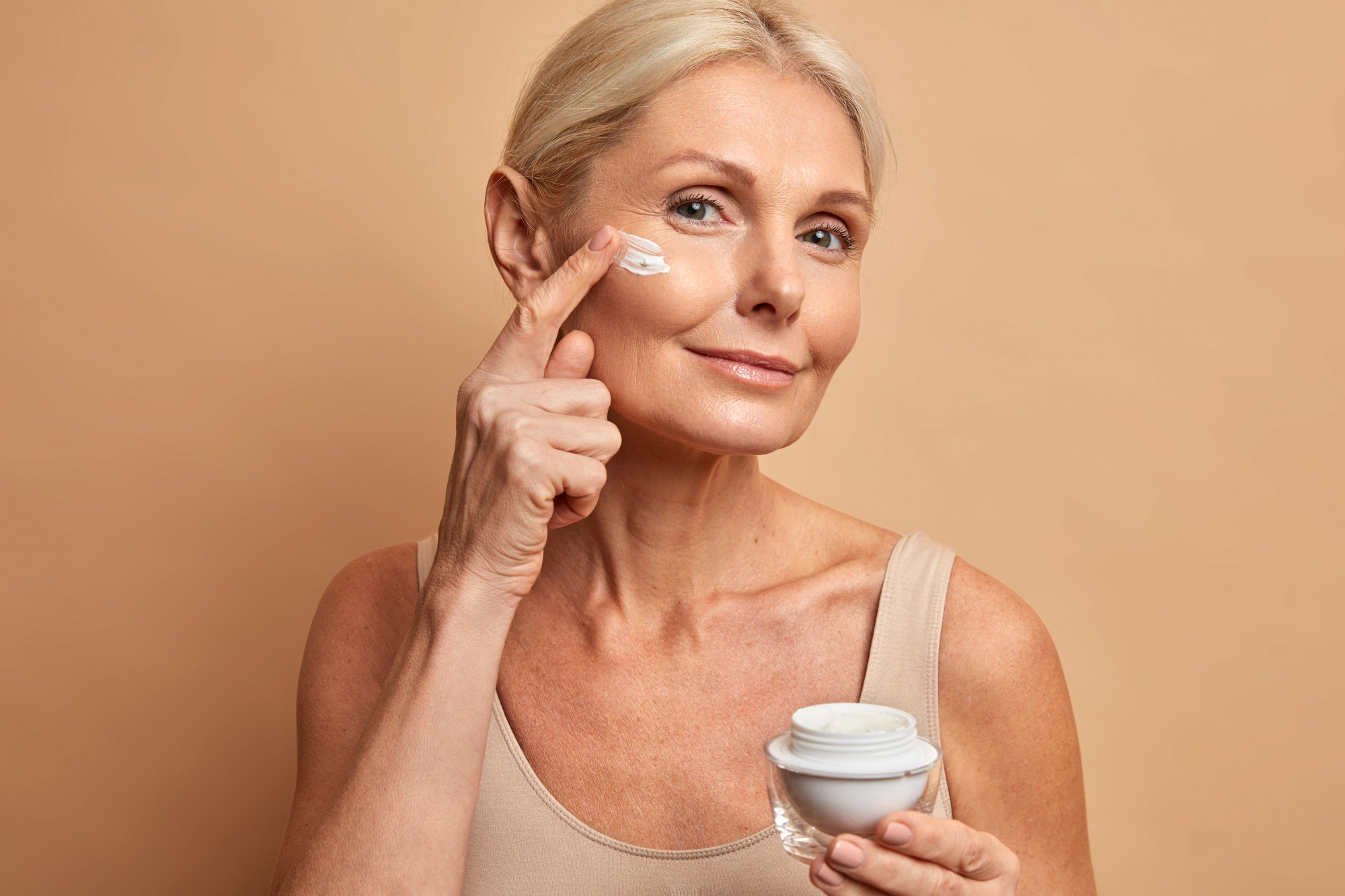 Anti-Aging.jpg Eine Frau mit blondem Haar trägt Gesichtspflegecreme auf ihre Wange auf, während sie einen Cremetiegel in der Hand hält.