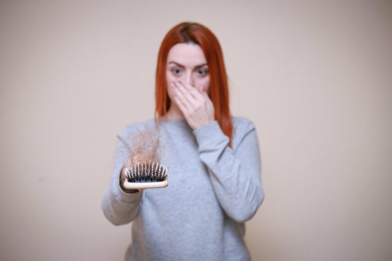 Haarausfall.jpg Frau mit rotem Haar hält eine Bürste mit Haaren in der Hand und zeigt einen überraschten Gesichtsausdruck.