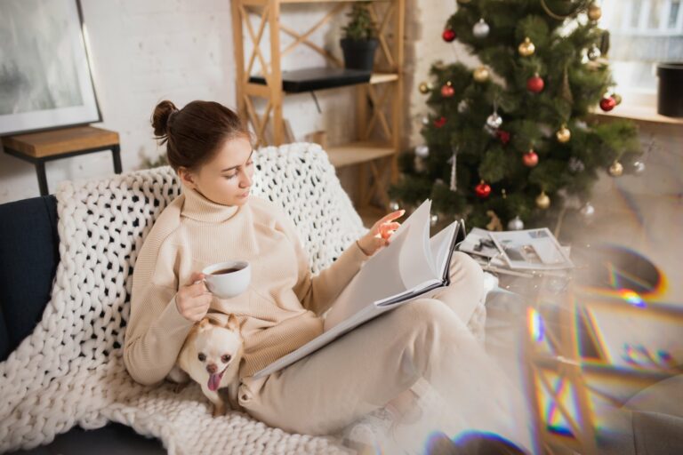 Eine Frau in bequemer Kleidung sitzt auf einem Sofa, liest in einem Buch und hält eine Tasse Kaffee, während ein Hund bei ihr sitzt.