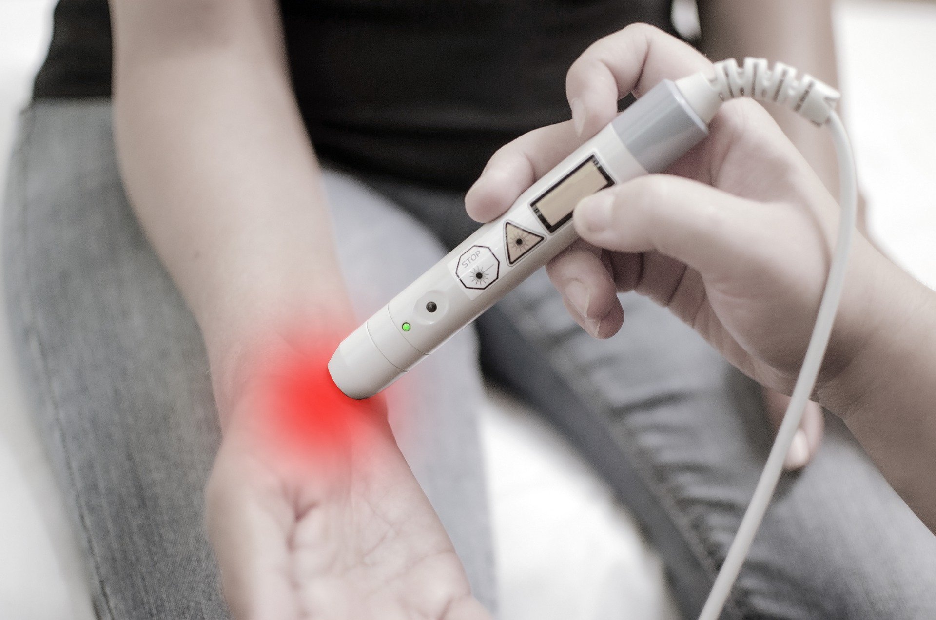 Karpaltunnelsyndrom.jpg Eine Hand hält ein medizinisches Gerät, das rotes Licht auf den Arm einer Person projiziert.