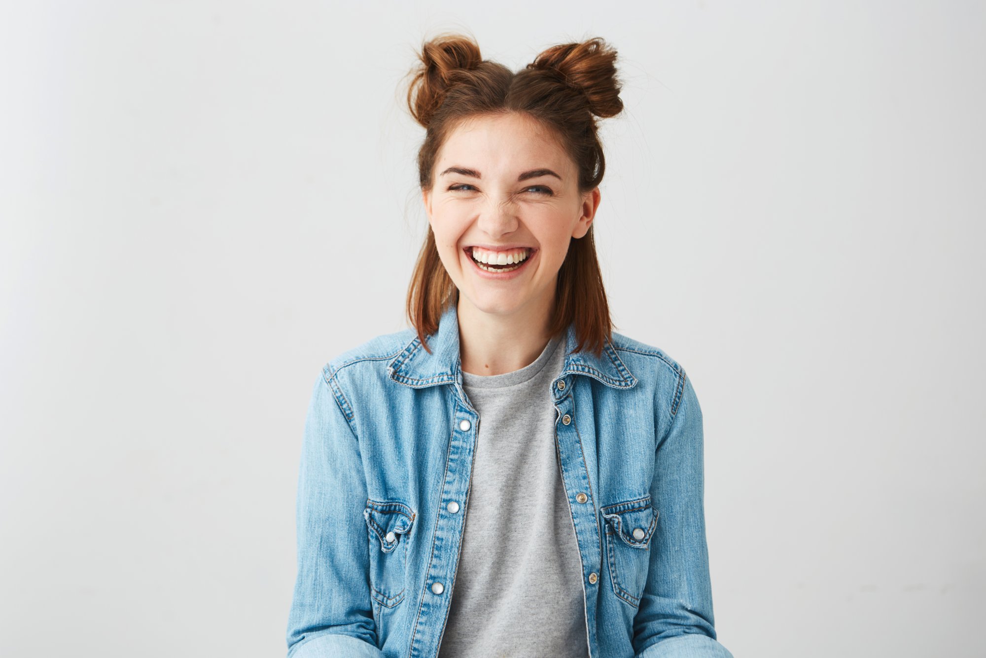Lachen.jpg Lachende junge Frau mit Duttfrisur, trägt eine Jeansjacke über einem grauen T-Shirt, vor einem hellen Hintergrund.