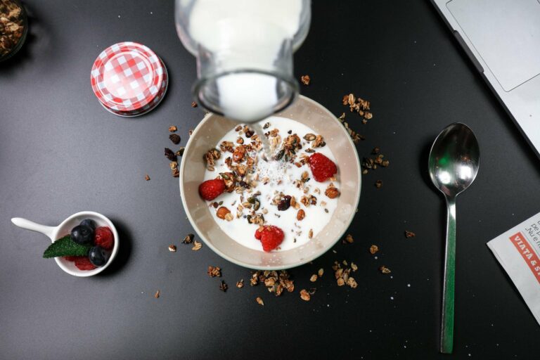 Ein Frühstücksgericht mit Joghurt, Granola und Himbeeren, während Milch darüber gegossen wird. Ein Löffel und eine Schale mit Beeren sind sichtbar.