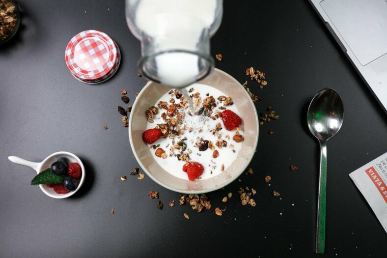 Ein Frühstücksgericht mit Joghurt, Granola und Himbeeren, während Milch darüber gegossen wird. Ein Löffel und eine Schale mit Beeren sind sichtbar.