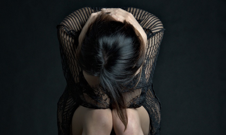 Eine sitzende Person in einer schwarzen Spitzenkleidung, die den Kopf mit beiden Händen umschließt und sich nach vorne beugt.