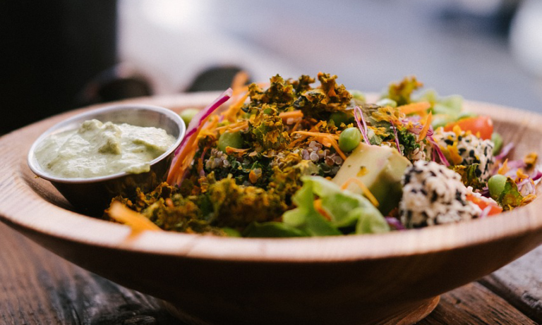 Bowl mit buntem Salat, darunter Quinoa, Karotten, Grünzeug und einer kleinen Schale Dressing.