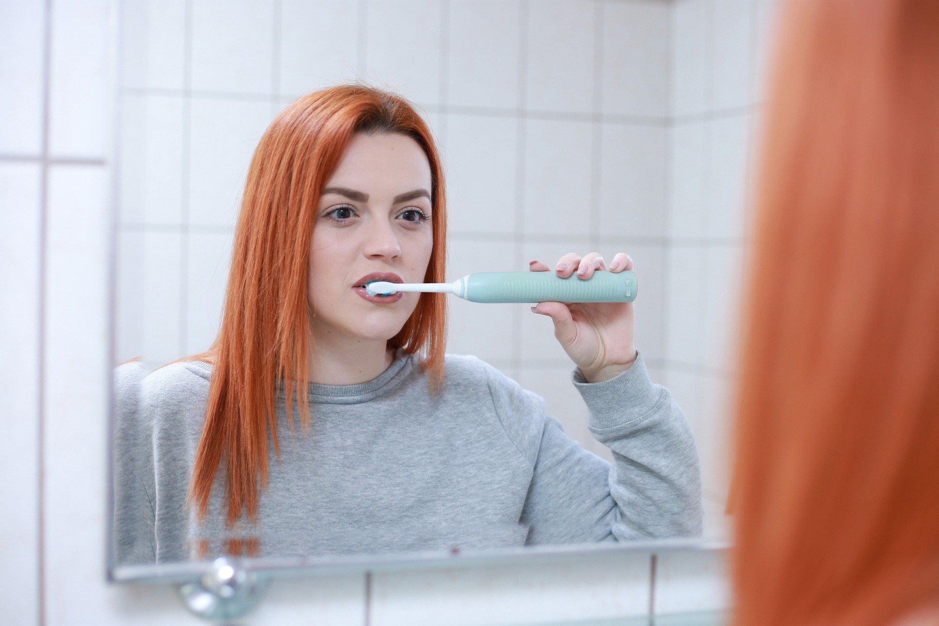 Mundhygiene.jpg Frau mit langen, roten Haaren putzt sich mit einer elektrischen Zahnbürste im Badezimmer die Zähne.