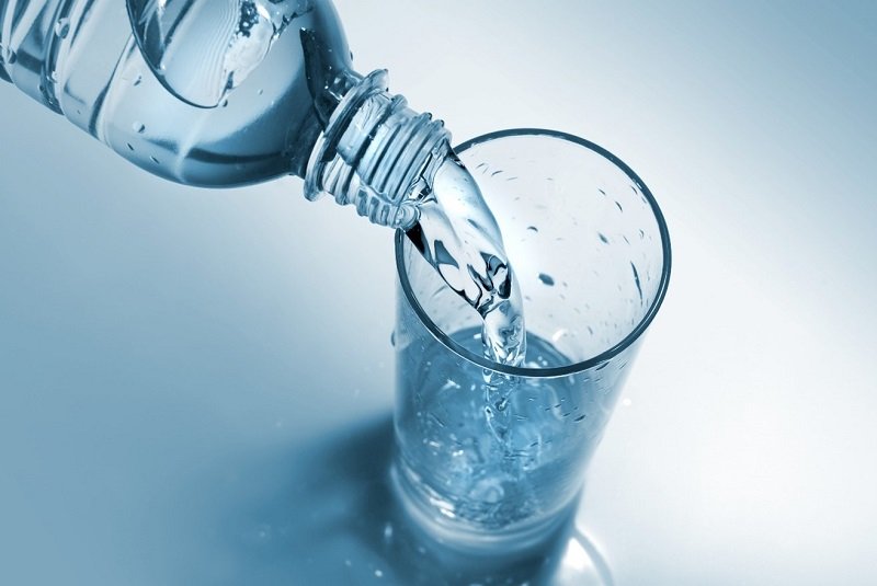 Wasserglas_WEB.jpg Eine Wasserflasche schüttet frisches Wasser in ein Glas, das auf einer hellen, blauen Oberfläche steht.
