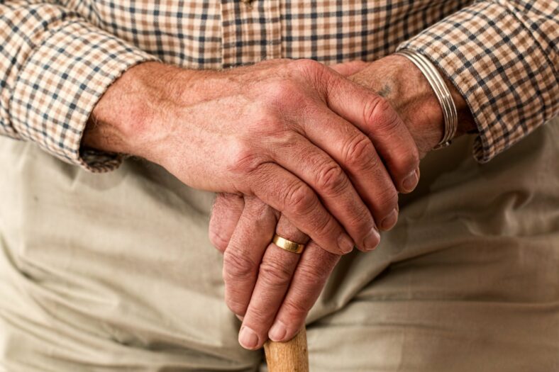 arthrose.jpg Faltenreiche Hände mit Ringen, die eine Holzstange halten, hinter einem karierten Hemd sichtbar.