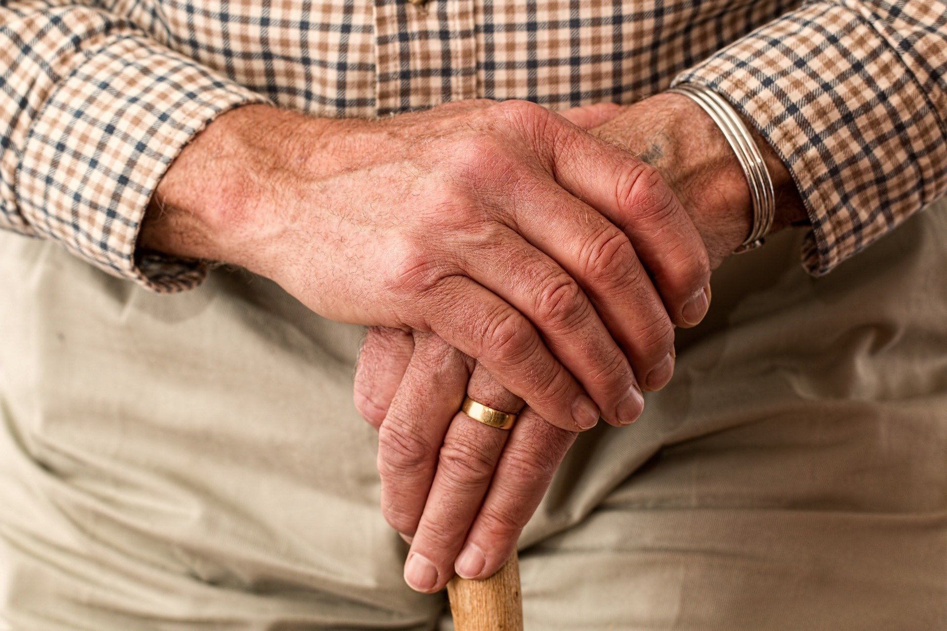 arthrose.jpg Faltenreiche Hände mit Ringen, die eine Holzstange halten, hinter einem karierten Hemd sichtbar.