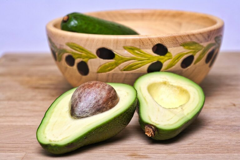 Eine halbierte Avocado mit einem großen Kern, daneben eine ganze Avocado und eine dekorative Holzschüssel im Hintergrund.