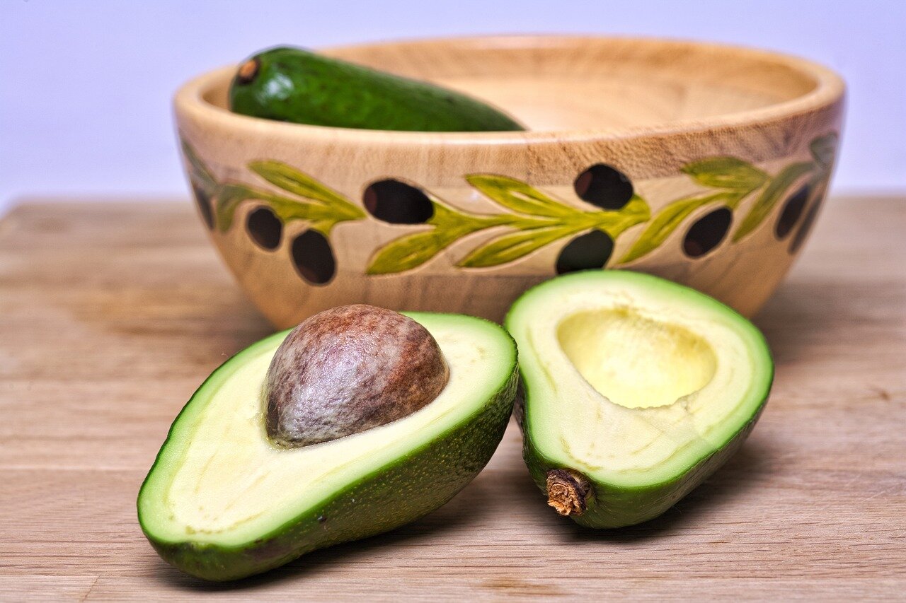 avocados.jpg Eine halbierte Avocado mit einem großen Kern, daneben eine ganze Avocado und eine dekorative Holzschüssel im Hintergrund.