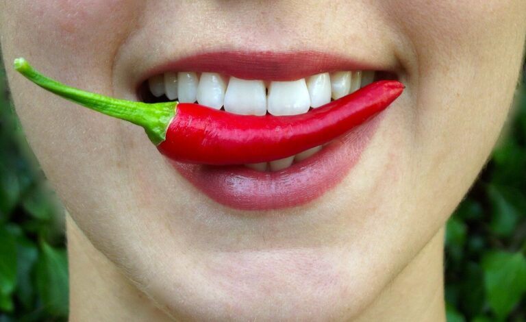 Eine Person mit einem roten Chili in den Lippen, lächelnd vor einem grünen Hintergrund.