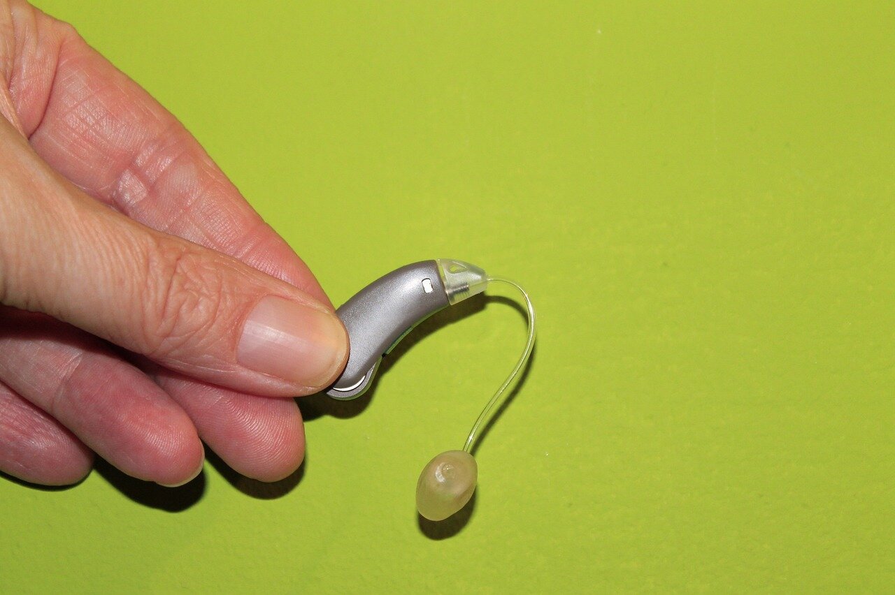 hoergeraet.jpg Eine Hand hält ein modernes Hörgerät mit einem durchsichtigen Schlauch und Ohrstück vor einem grünen Hintergrund.
