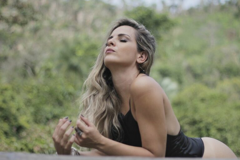 Eine Frau in einem schwarzen Oberteil liegt im Freien und blickt nachdenklich in die Ferne, Hintergrund mit grünem Bewuchs.