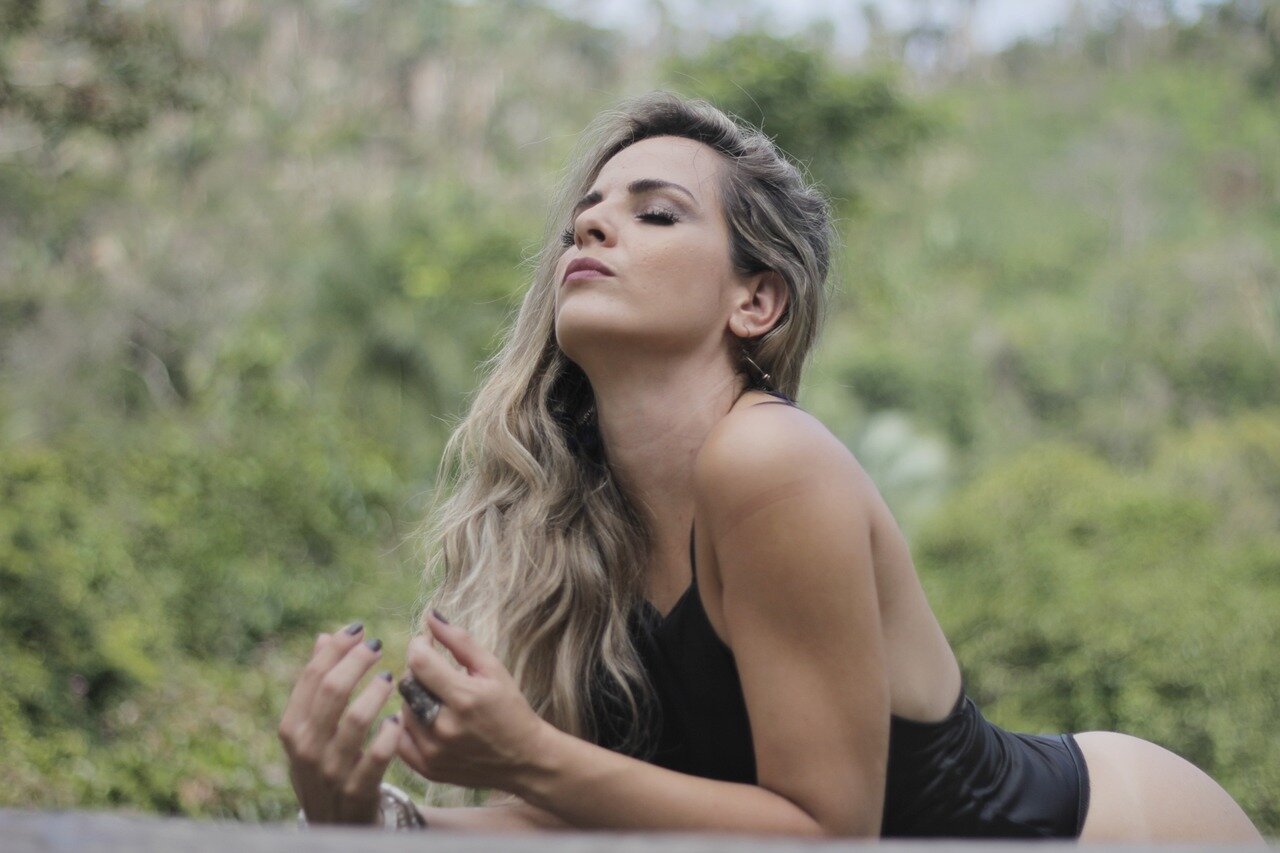 kraft-der-atmung.jpg Eine Frau in einem schwarzen Oberteil liegt im Freien und blickt nachdenklich in die Ferne, Hintergrund mit grünem Bewuchs.