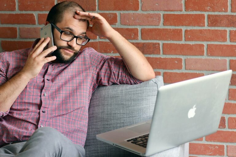 Mann mit Brille sitzt auf einem Sofa, telefoniert und schaut besorgt auf einen Laptop.