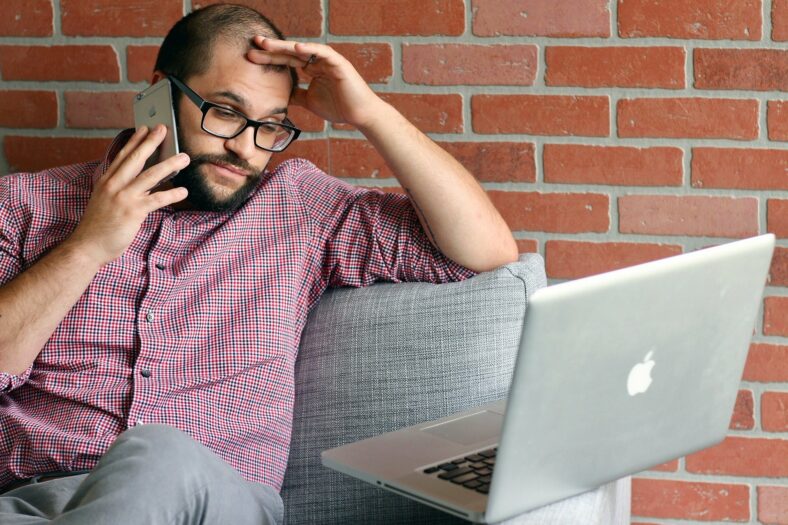 stress-reduktion.jpg Mann mit Brille sitzt auf einem Sofa, telefoniert und schaut besorgt auf einen Laptop.