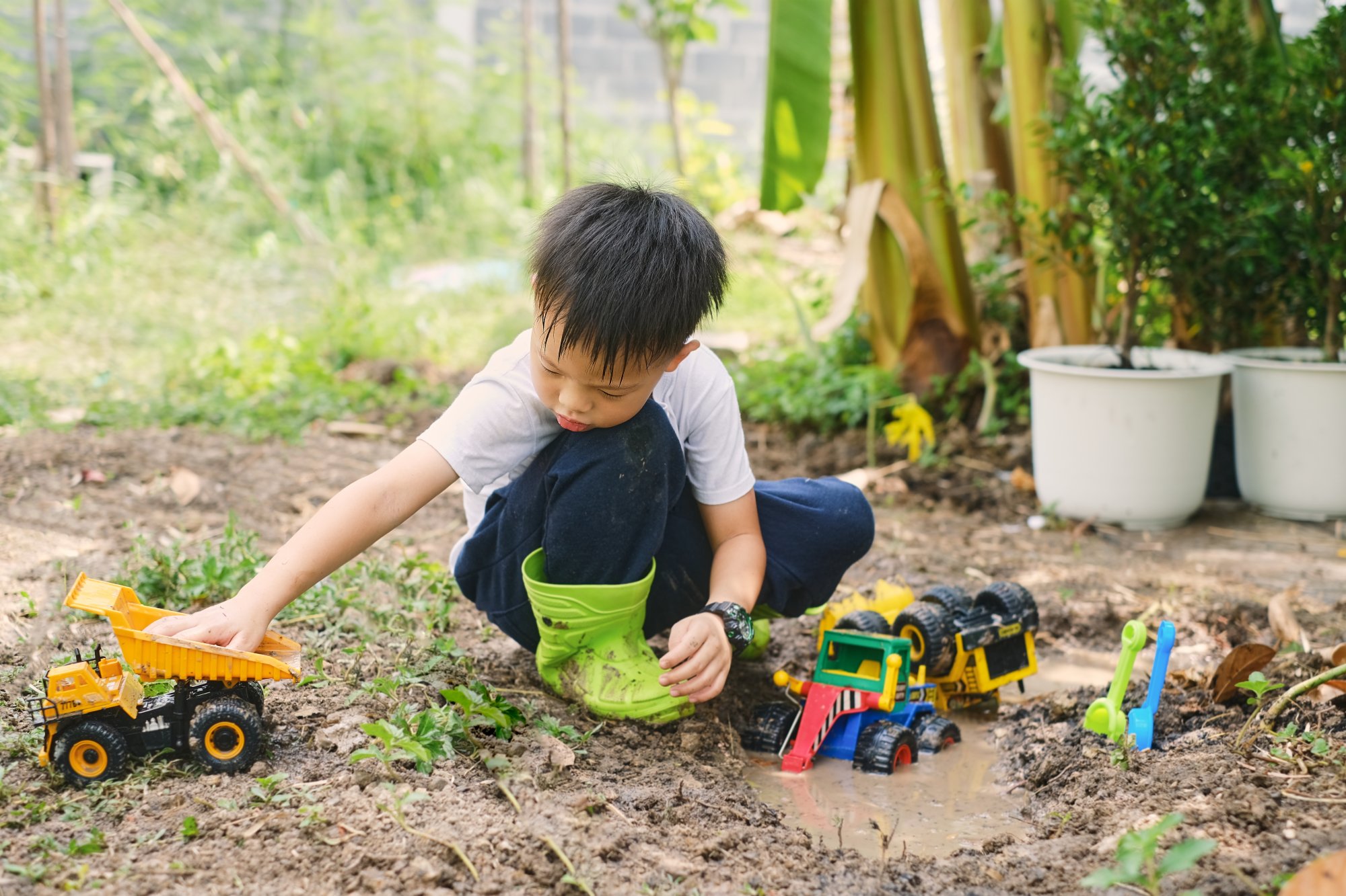 wie-viel-dreck-ist-gesund.jpg Ein Junge im Garten spielt mit verschiedenen Spielzeugfahrzeugen, darunter ein Lkw und ein Bagger, während er in matschigem Boden sitzt.