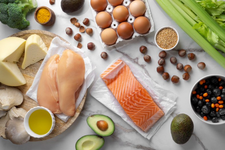 Proteinreiche_Ernaehrung.png Verschiedene Lebensmittel wie Hähnchenbrust, Lachs, Käse, Eier, Avocado und Gemüse auf einem Tisch.