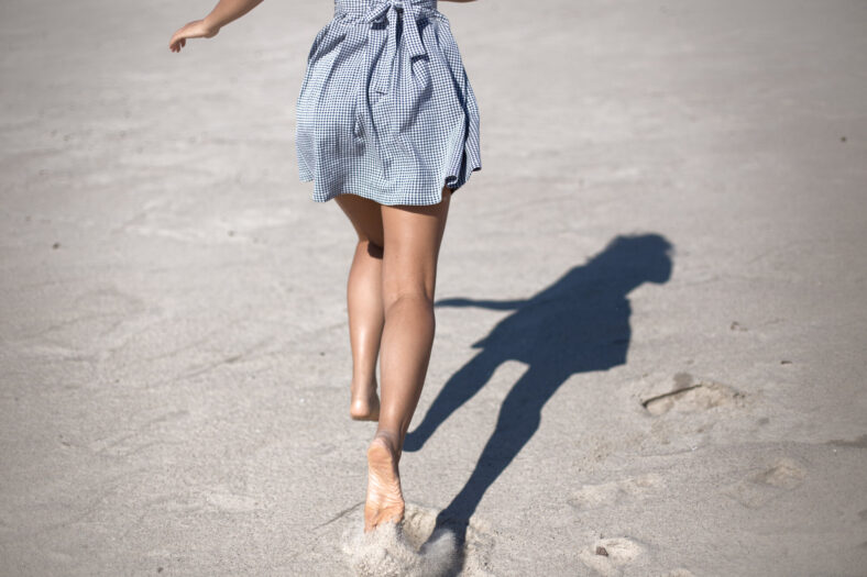 Eine Person in einem karierten Kleid läuft barfuß über einen sandigen Strand, während ihr Schatten sichtbar ist.
