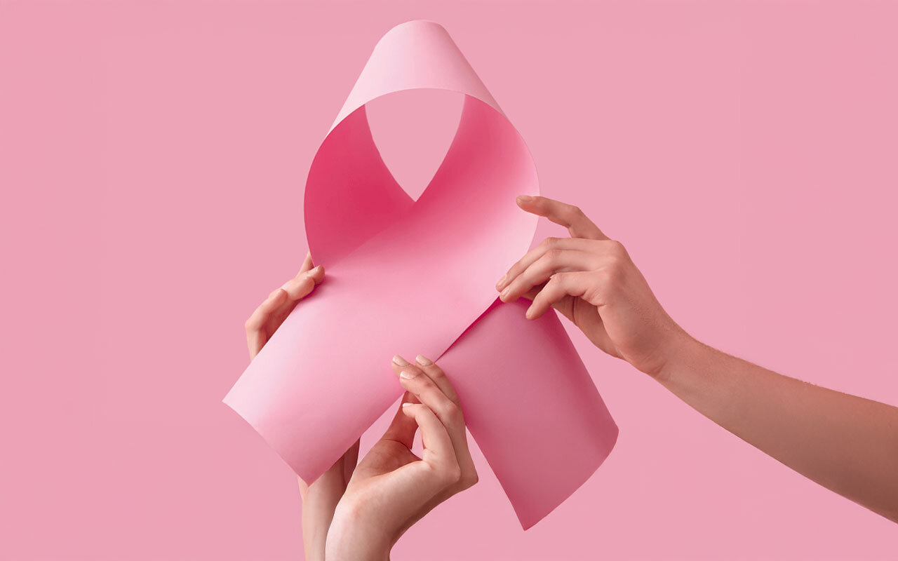Welt-Brustkrebs-Tag.jpg Zwei Hände halten ein rosa Papier, das zu einer Schleife geformt ist, vor einem pinken Hintergrund.